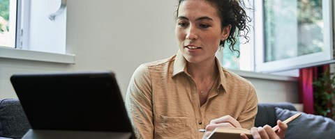 kvinne som sitter i sofaen og tar notater foran en pc-skjerm.